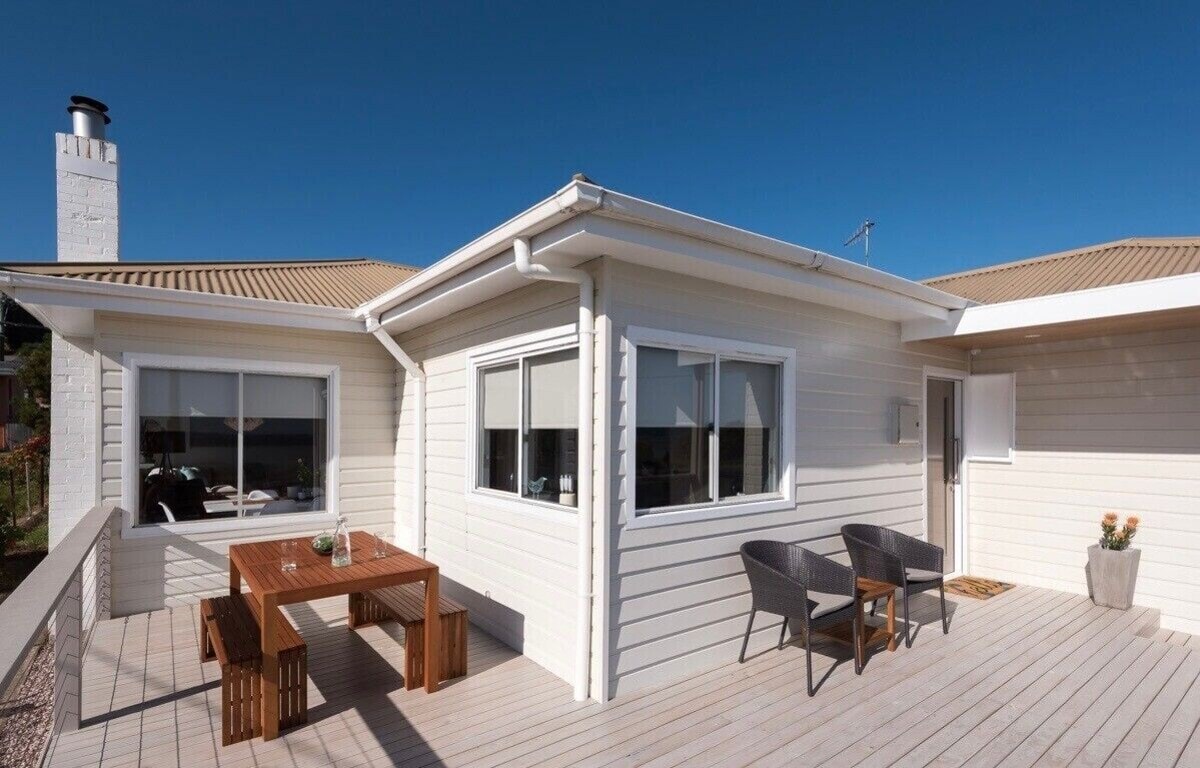 Rear deck with timber outdoor dining table, bench seats and rattan chairs under blue sky