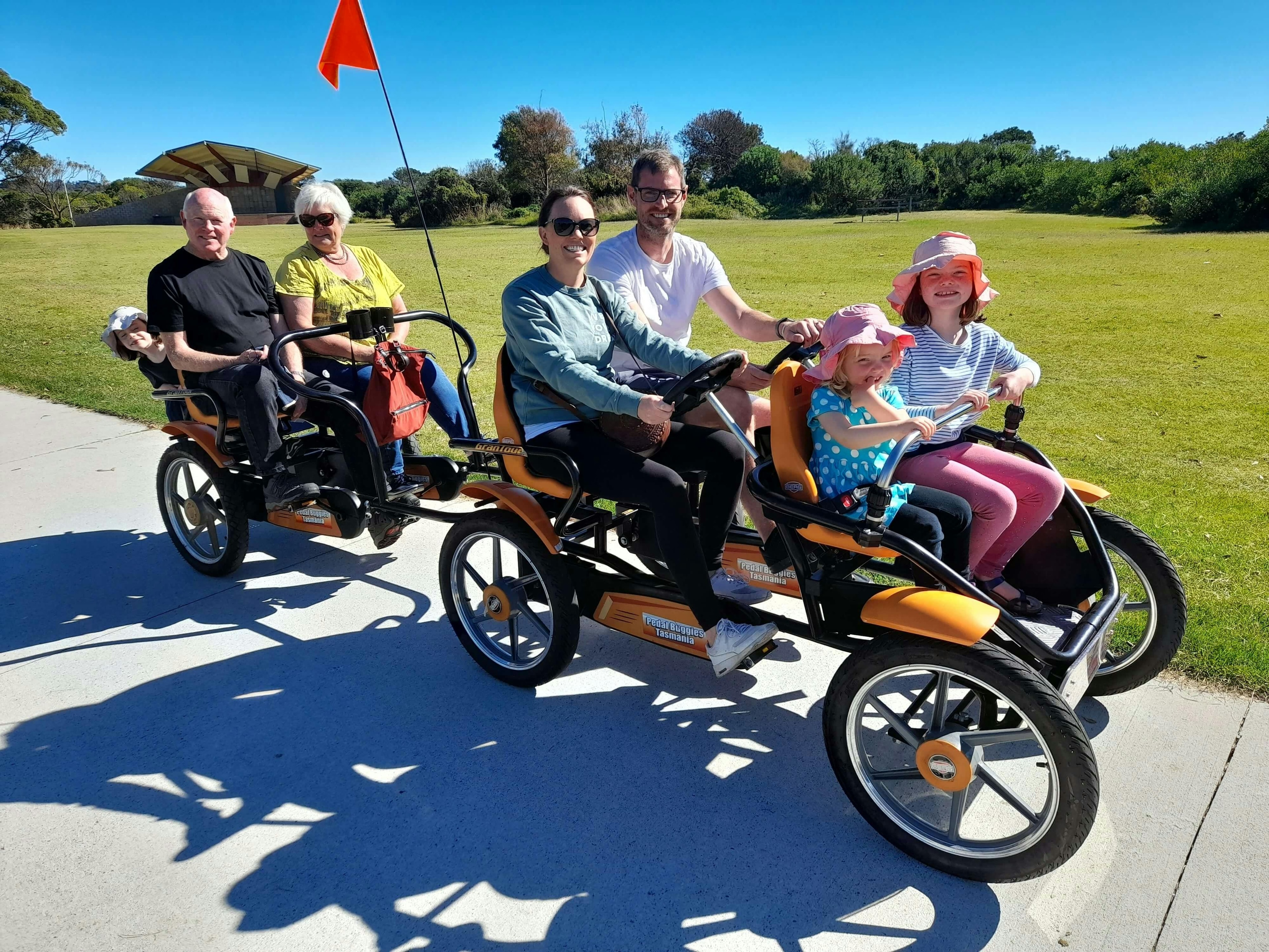 Family riding orange Pedal Buggies Tasmania four-wheeled bikes along a coastal path on a sunny day